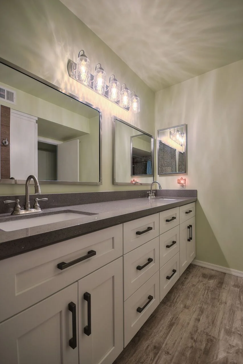 White bathroom vanity with dark countertop, dual sinks, mirrors, and vanity lighting