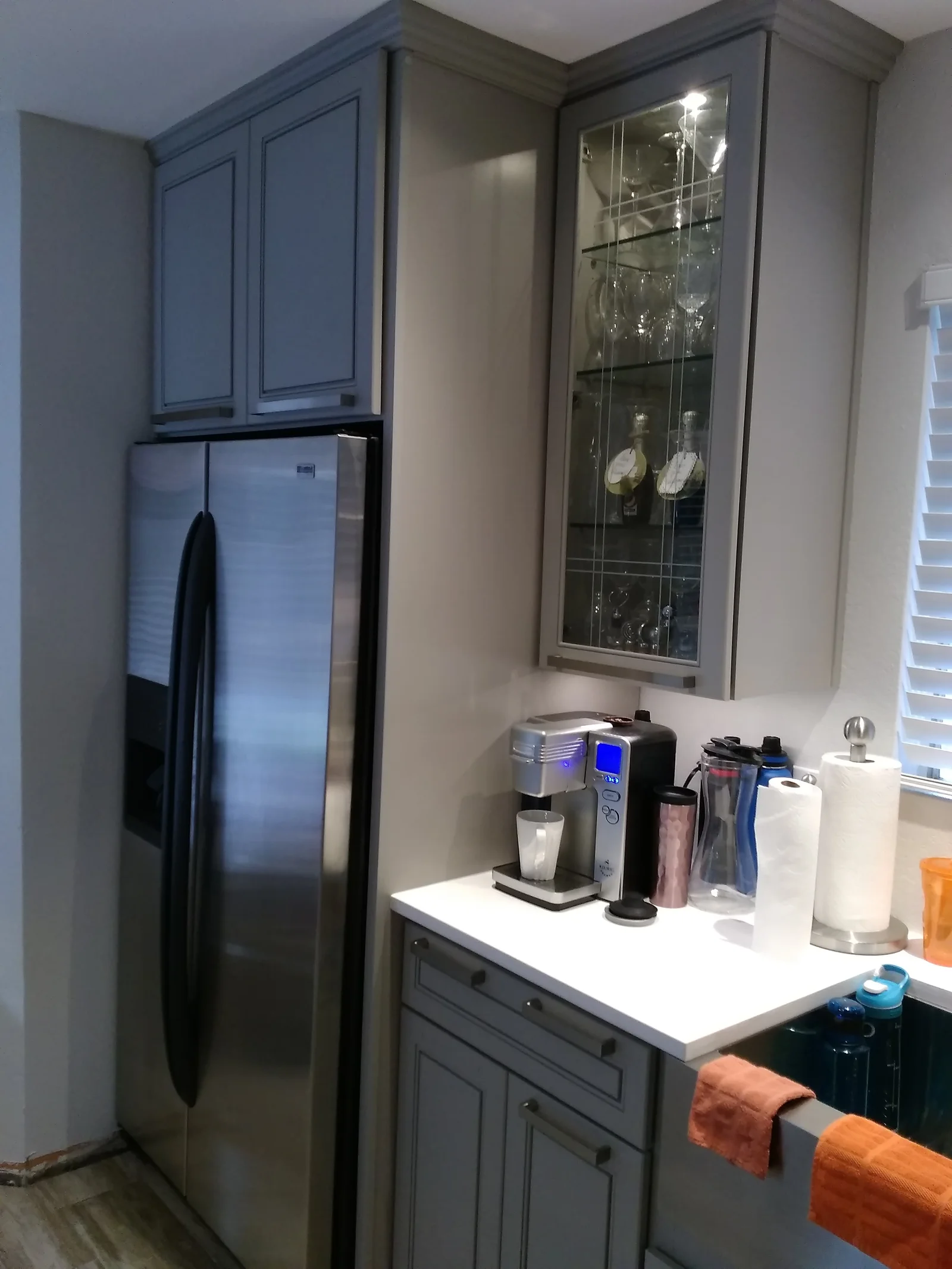 Gray kitchen cabinets with white quartz countertop, stainless steel refrigerator, and glass display cabinet